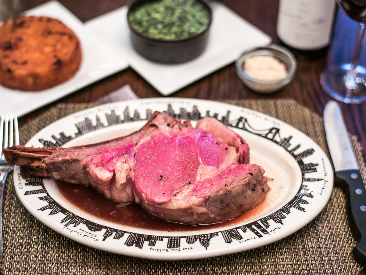 A delectable slice of rare prime rib adorning an elegant oval plate, accompanied by a fork, a steak knife, and an assortment of small side plates and a wine glass.