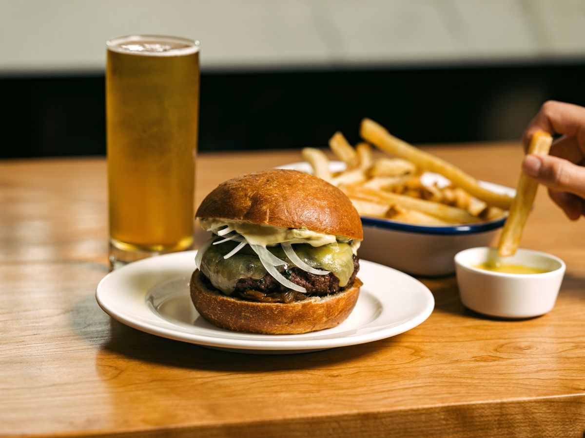 A hand submerges a french fry into a small white dish of sauce next to a burger sitting on a plate.