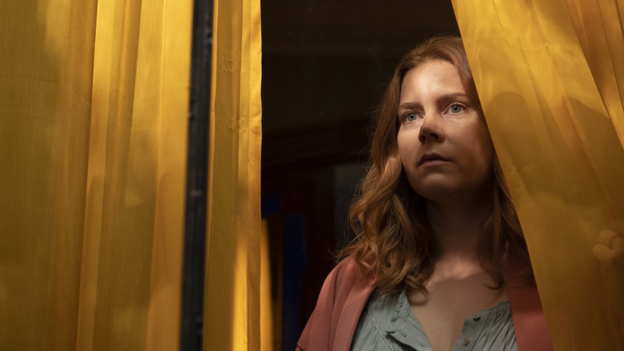 A woman gazes out of the window, her visage surrounded by drapes.