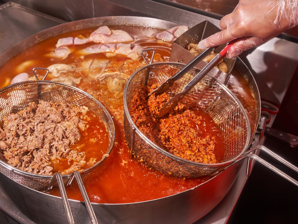 The image exhibits a gloved hand holding a sieve containing crumbly red meat over a vat of orange fat and oil, alongside other meats.